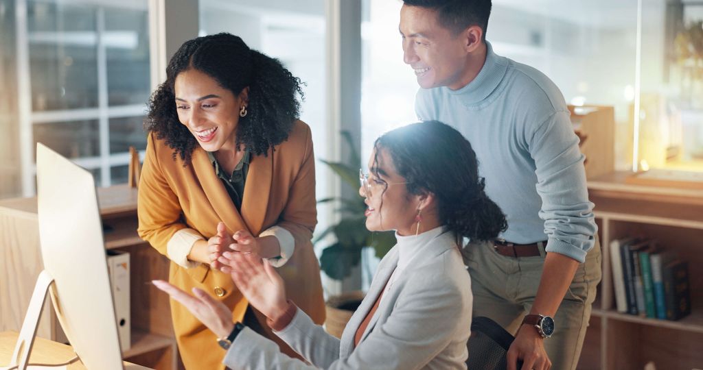 Drei Personen arbeiten gemeinsam an einem Schreibtisch. Sie lächeln und blicken auf einen Computerbildschirm, während eine Person gestikuliert. Die Gruppe scheint in ein positives Gespräch vertieft zu sein, während sie in einem modernen, hellen Büro arbeiten.